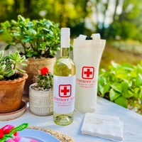 White wine bottle with 'Thirst Aid' label on a table outdoors with plants in the background and placed beside a matching  "Thirst Aid" matching wine tote. This fun label is perfect for birthdays, girls' nights, bachelorettes, or as a cute nursing gift.