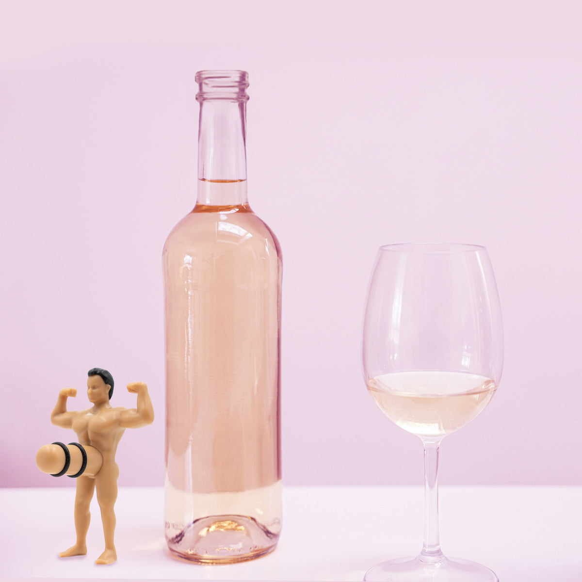 Bottle of pink wine with a glass and a small figurine wine bottle stopper on a light pink background
