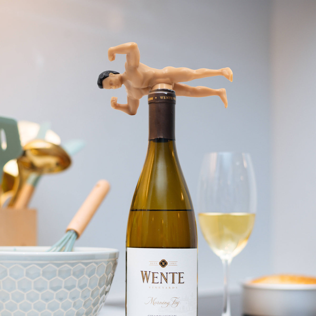 White wine bottle with a humorous bottle stopper shaped like a man with the dick as the wine stopper, set against a casual dining background.