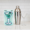 A stainless steel cocktail shaker and a clear martini glass on a white countertop, next to a packaged kitchen sponge with a martini glass illustration and the text 'TINI BIT DirtY'.