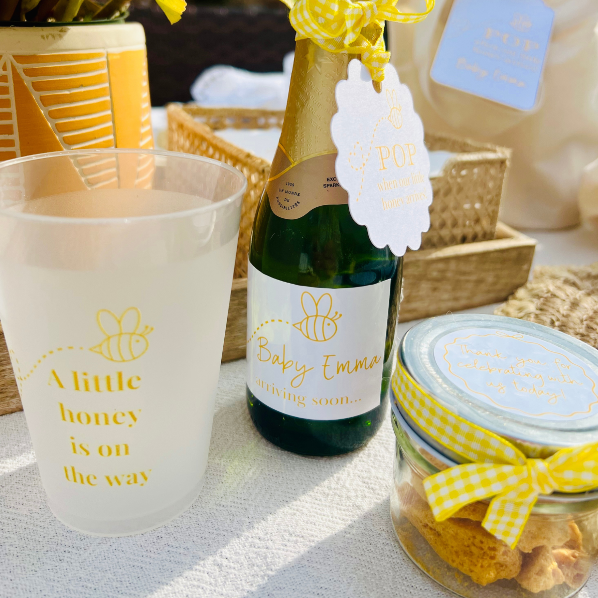Bottle of mini champagne for a bee-themed baby shower is shown alongside other party favors,  including a matching party cup and sweet jar. 
