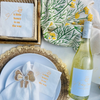 Table setting with white wine bottle, plates, and napkins with text on a light background. The napkins and wine bottle feature a matching bee-themed baby shower design. 