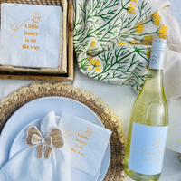 Table setting with white wine bottle, plates, and napkins with text on a light background. The napkins and wine bottle feature a matching bee-themed baby shower design. 