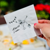 White cocktail napkin is held up against a garden background. The napkin features a girl in a martini glass beside text that reads "A tini bit older"