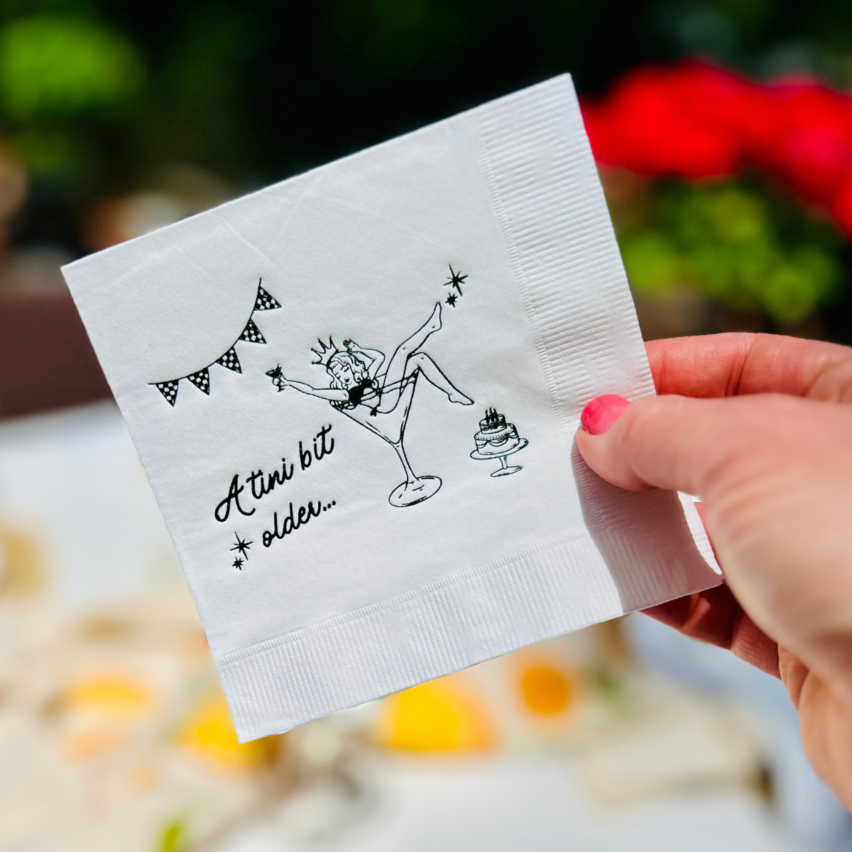 White cocktail napkin is held up against a garden background. The napkin features a girl in a martini glass beside text that reads "A tini bit older"