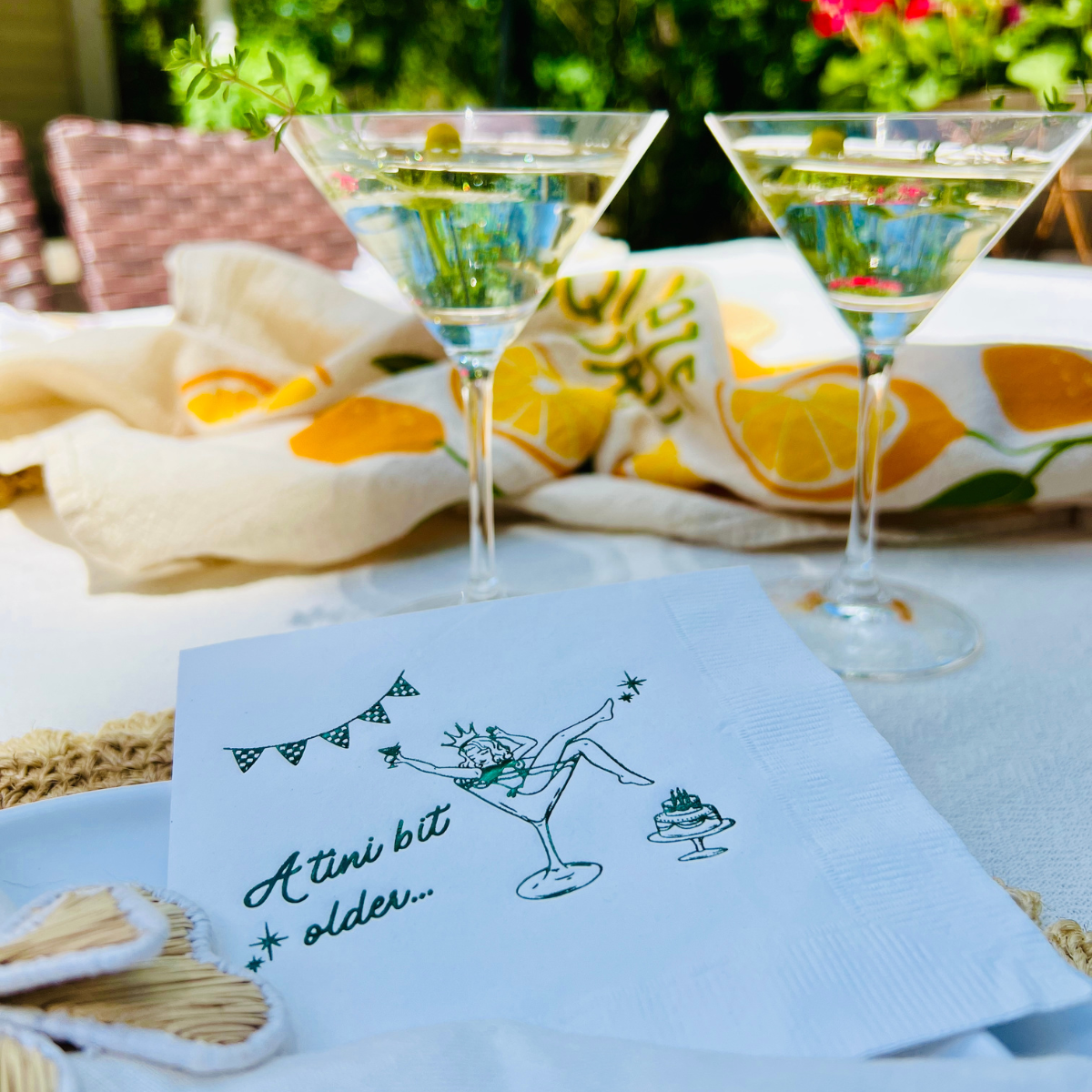 Two martini glasses with chamomile garnishes on a tablecloth with a cocktail napkin featuring text and graphics.