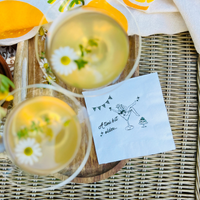 Two chamomile lemon martinis beside a white paper cocktail napkin that features a design of a woman in a martini glass and text that reads "A Tini Bit Older" for martini -themed birthday parties.