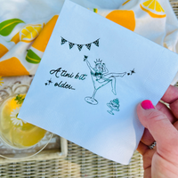 Napkin with a design and text held by a hand, with a colorful fabric background. The paper cocktail napkin features a design of a woman in a martini glass and text that reads "A tini Bit Older"