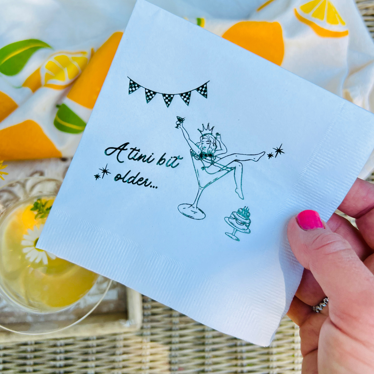 Napkin with a design and text held by a hand, with a colorful fabric background. The paper cocktail napkin features a design of a woman in a martini glass and text that reads "A tini Bit Older"