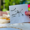 Napkin with a fun design of a woman in a martini glass, text 'Attini bit older', and a cake, held by a hand.