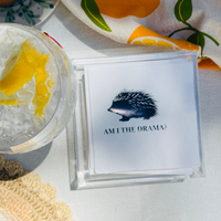 White cocktail napkin with hedgehog illustration and 'Am I the Drama?' text on a table with a drink and towel.
