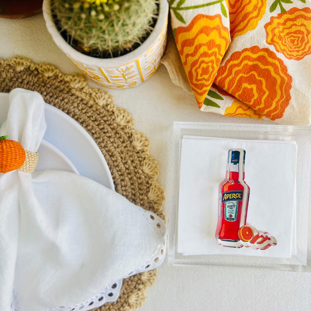Table setting with an acrylic napkin weight in the shape of an Aperol bottle is shown on a stack of white napkins. 