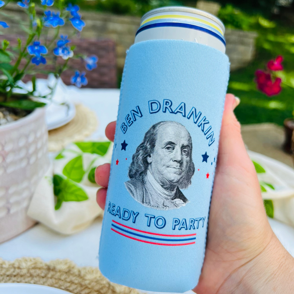 Koozie with Ben Franklin design held in a hand with a garden background