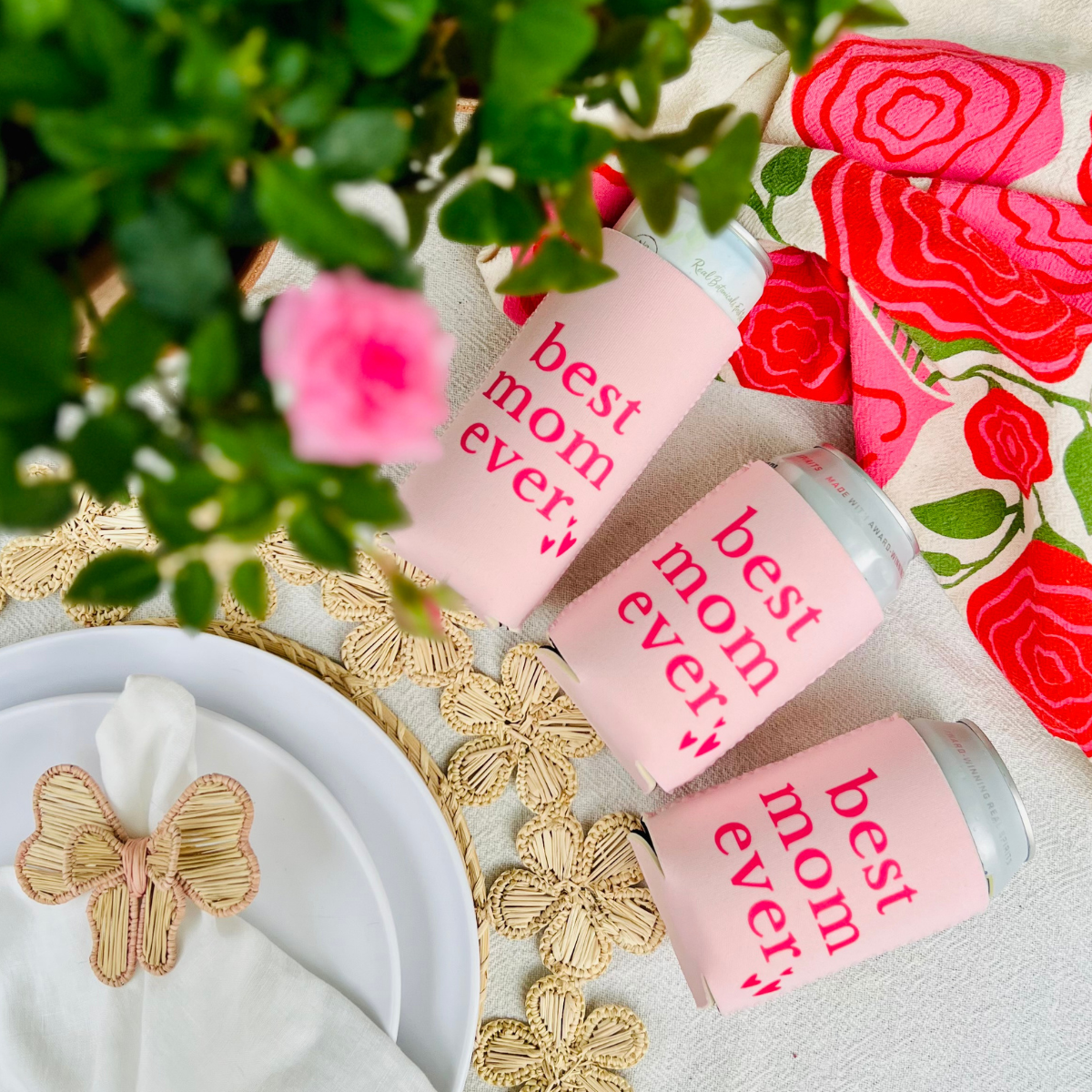 Three cans with can cooler sleeves that feature text that reads  'best mom ever' on a decorative surface with flowers and ribbons.