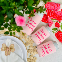 Three cans with can cooler sleeves that feature text that reads  'best mom ever' on a decorative surface with flowers and ribbons.