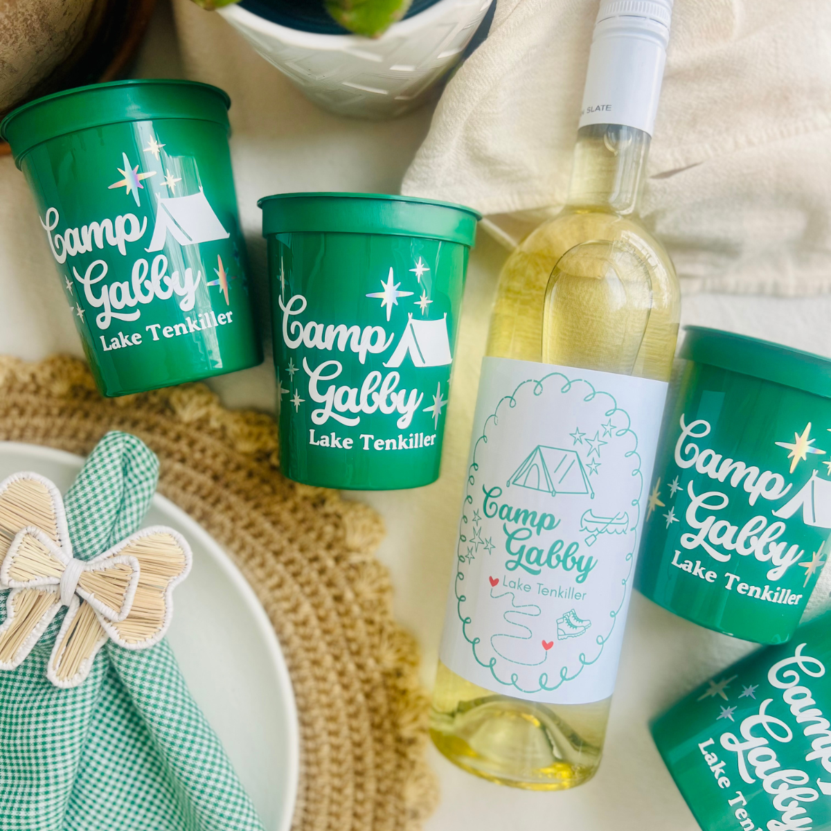 Green 'Camp Gabby' cups and a bottle of white wine with a matching wine label is shown on a table. 