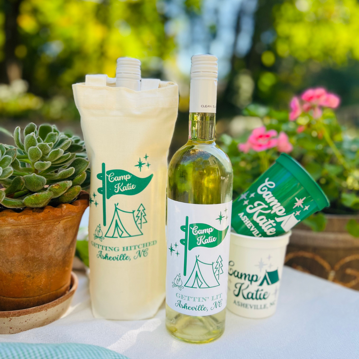 Camp Bachelorette themed wine label, matching wine bag, and customizable cups on a table in a nature setting.