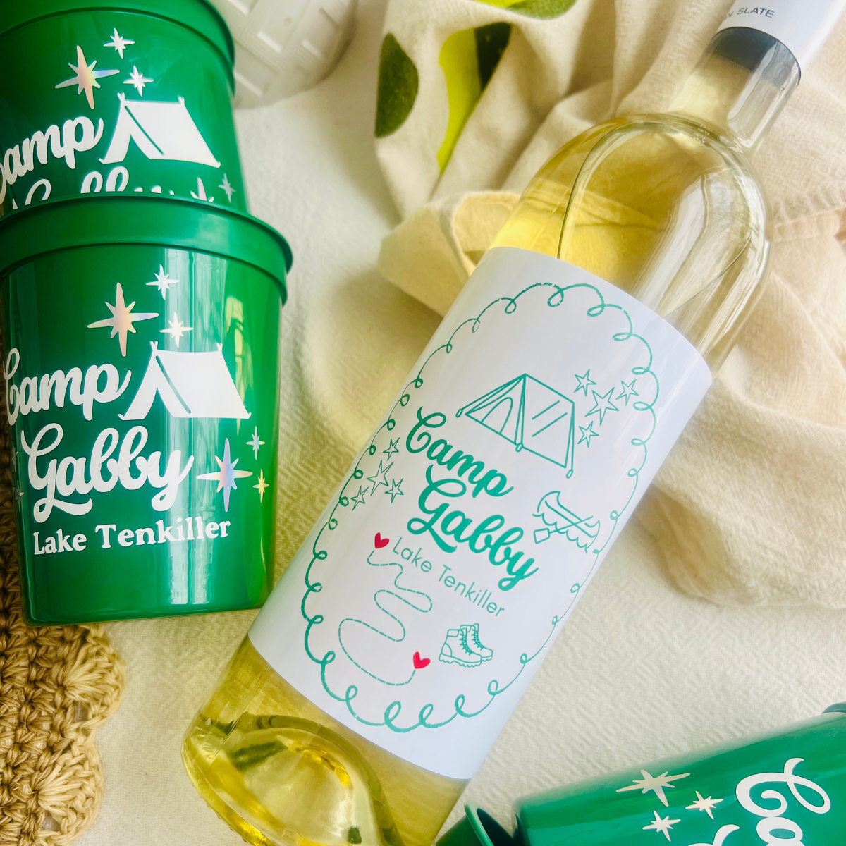 Green 'Camp Galby' cups and a bottle of wine with matching label on a beige fabric background.