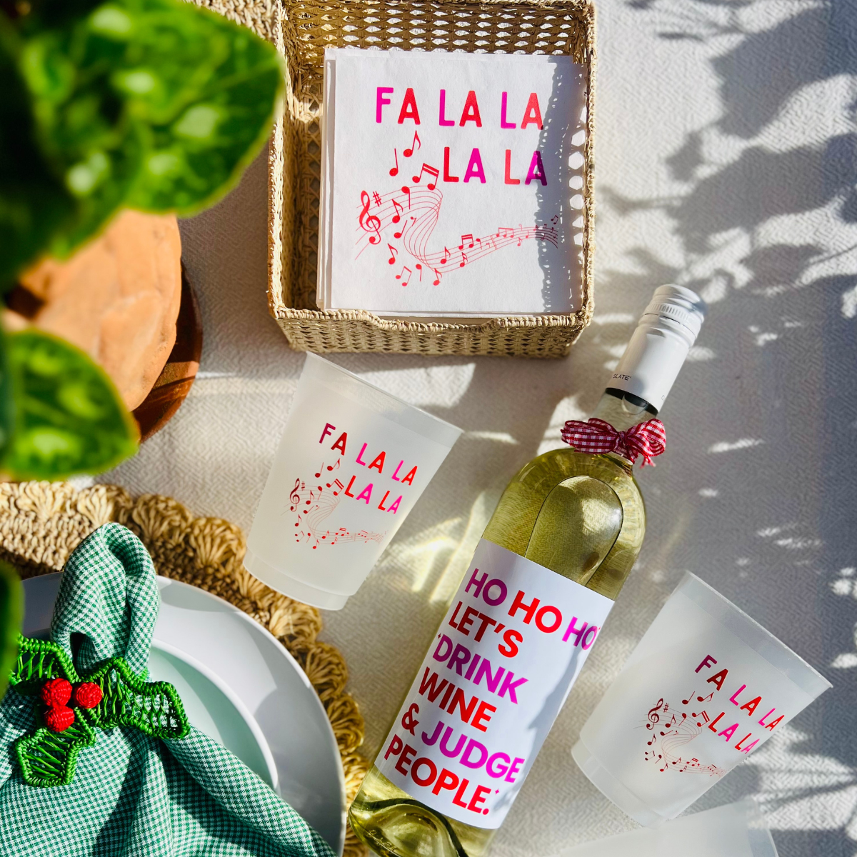 Overhead shot of a table setting with a bottle of white wine that features a fun wine label that reads "Ho Ho Ho Let's Drink Wine & Judge People" text in bold pink and red. The wine bottle is shown next to matching "Fa La La" Christmas napkins.