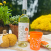 Wine bottle with "Fall In Love" Bachelorette Sunshine Daisy Wine Label shown on table beside fall décor and matching "Fall In Love" Stadium Cup