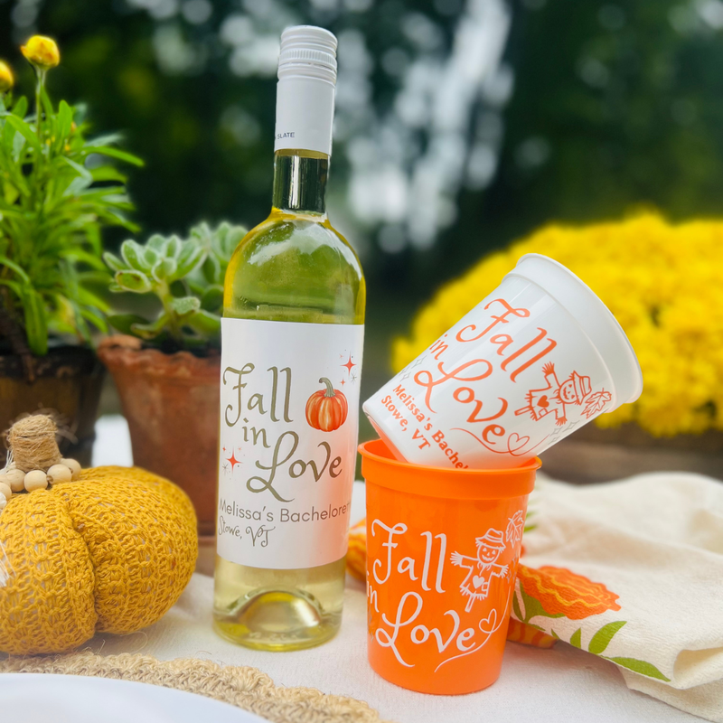 Fall tablescape showing a wine bottle featuring a Sunshine Daisy "Fall In Love" Wine Label beside two stacked "Fall In Love" Customizable Stadium Cups