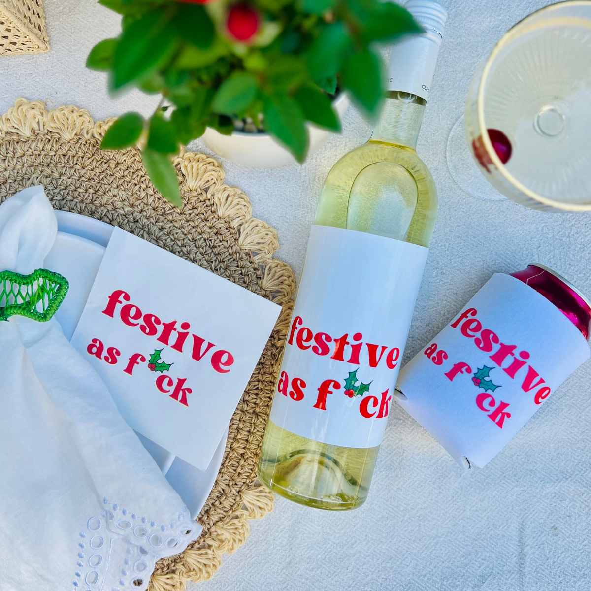 Bottle of white wine with a humorous holiday label features the fun text "festive as f*ck"  accompanied by a matching can cooler and napkin on a holiday table.