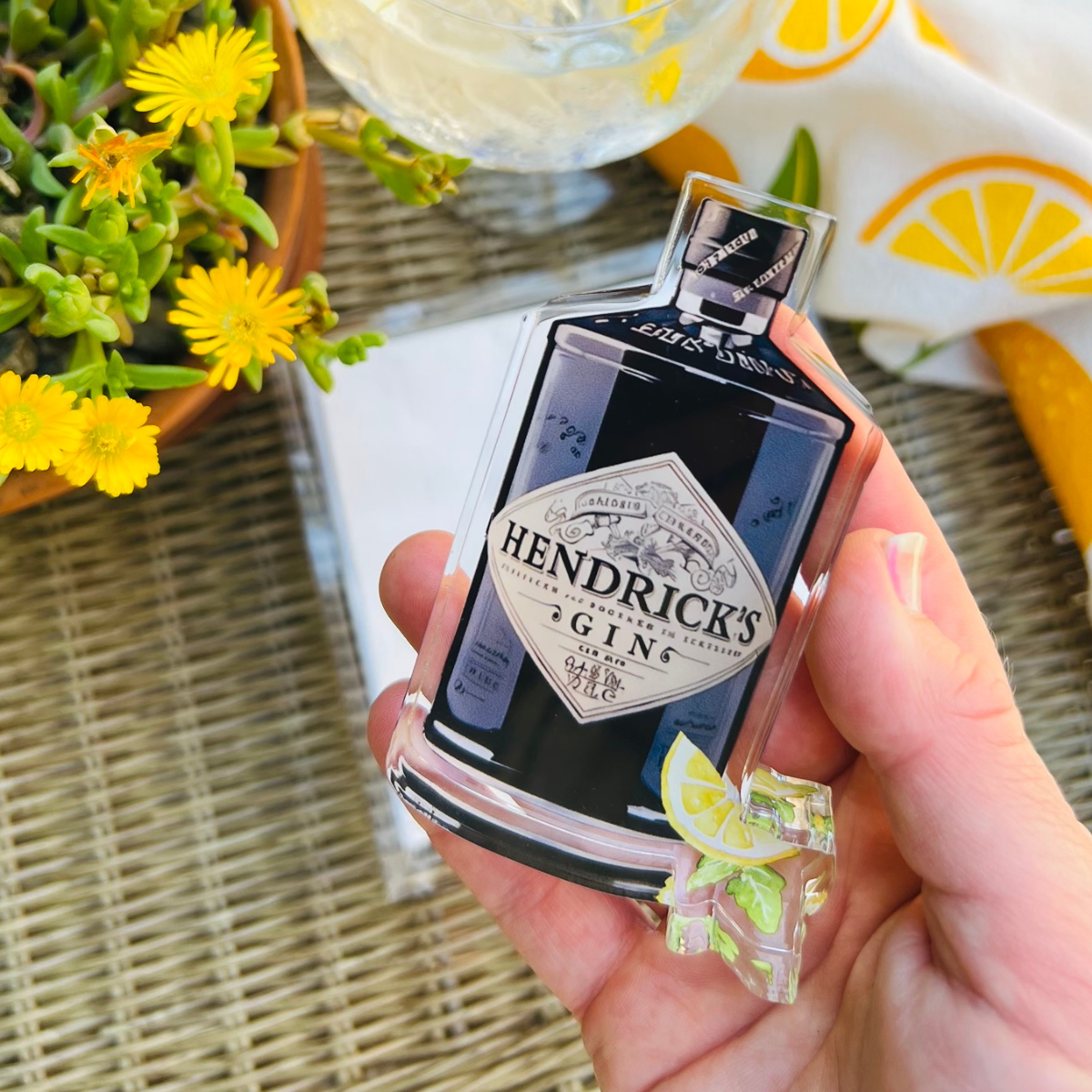 Hand holding a Hendrick's Gin napkin weight bottle in an outdoor setting with plants and tablecloth.