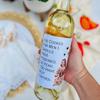 Wine bottle is held in an outdoor setting. The wine bottle has a wine label that features an illustration of a woman with a pot beside text that reads "I've cooked for me I should have poisoned, so yeah, I do have self control."