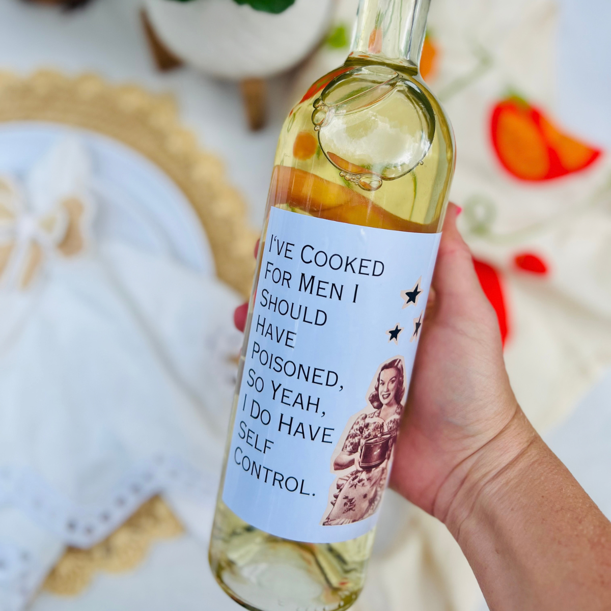 Wine bottle is held in an outdoor setting. The wine bottle has a wine label that features an illustration of a woman with a pot beside text that reads "I've cooked for me I should have poisoned, so yeah, I do have self control."