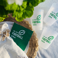 Football tail gaiting cups and napkins are shown on a table. The frost flex cups and green paper napkin feature a matching design that has a football graphic and text that reads "In My Football Era"