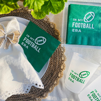 Green 'In My Football Era' napkins and cups on a textured surface with a decorative bow.