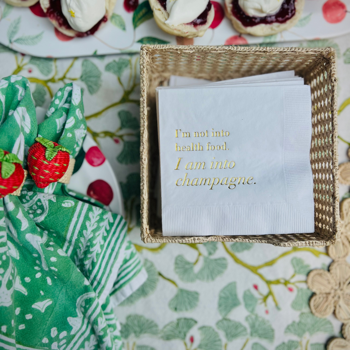 White paper cocktail napkin is shown in a raffia napkin holder. The napkin features gold foil text that reads "I'm not into health food, I am into champagne"