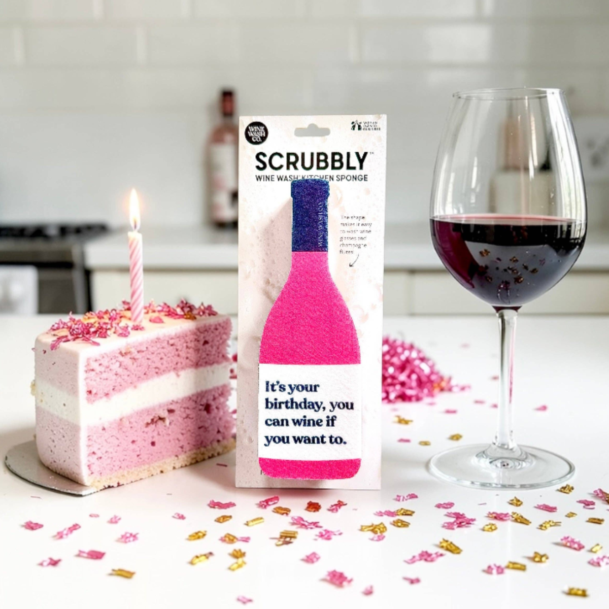 A boxed Scrubbly wine marker sponge stands on a kitchen counter beside a pink birthday cake slice with a candle and a glass of red wine, with confetti scattered on the surface.