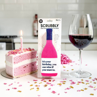 A boxed Scrubbly wine marker sponge stands on a kitchen counter beside a pink birthday cake slice with a candle and a glass of red wine, with confetti scattered on the surface.