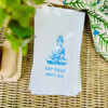 White pindot paper guest towel with blue Buddha illustration and text on a woven surface with greenery.