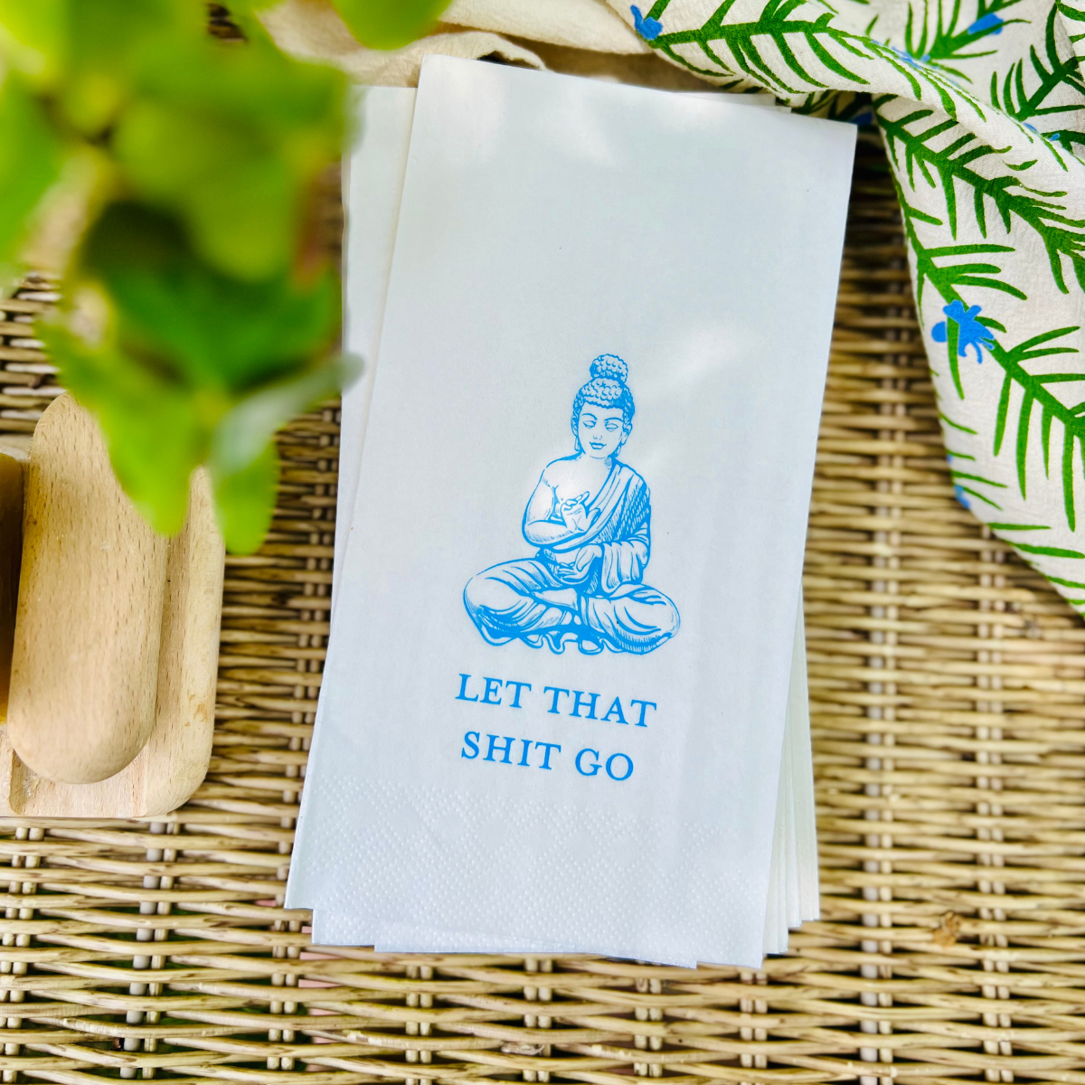 White pindot paper guest towel with blue Buddha illustration and text on a woven surface with greenery.