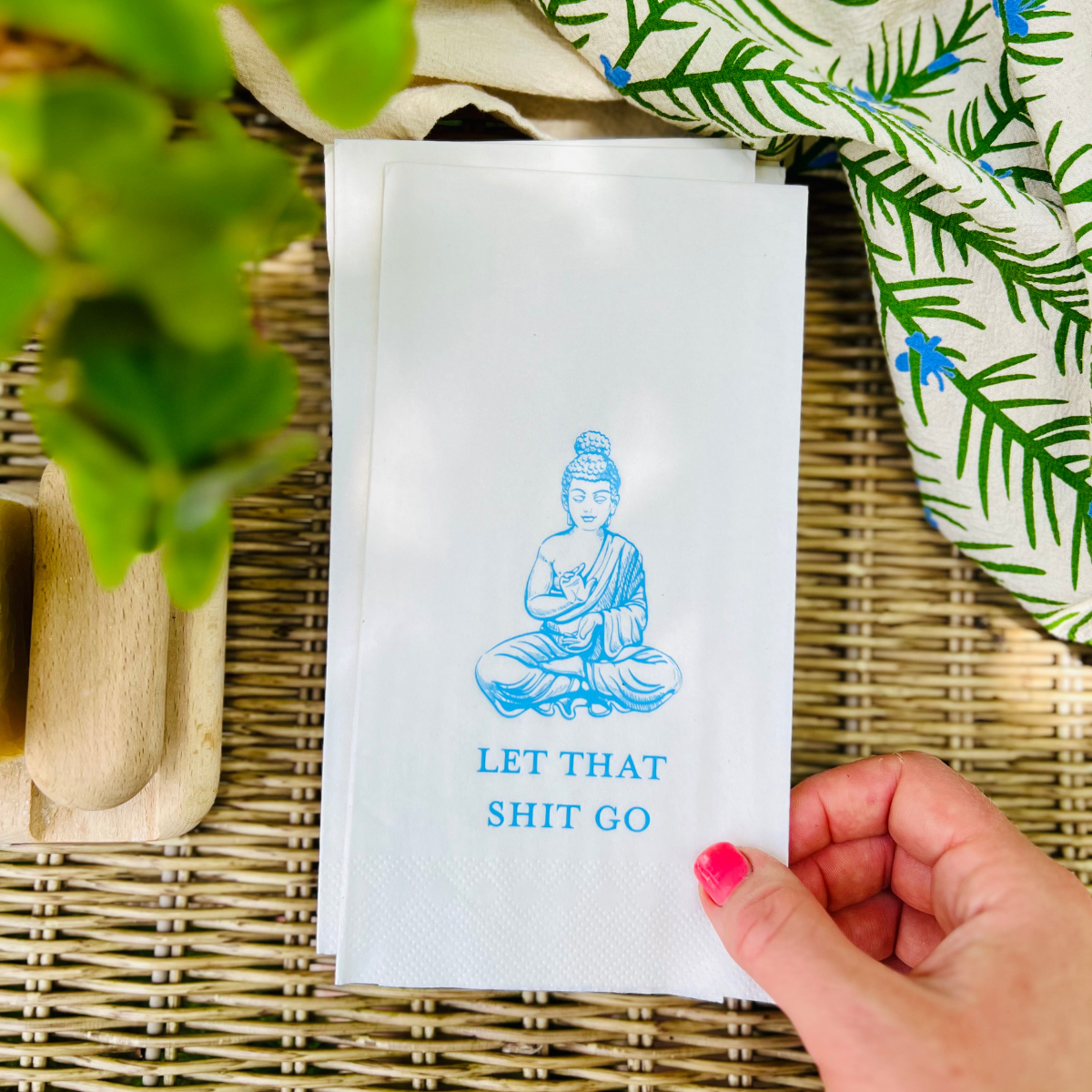 Hand holding a white paper guest towel with a blue Buddha illustration and text on a woven surface.