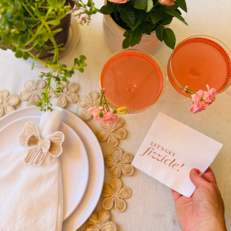 Anniversary, bridal shower, or birthday napkin, this white cocktail napkin features the words "Let's Get Fizzicle" in shiny rose gold font. The napkin is styled alongside a pretty placesetting and cocktails in coupe glasses.