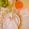 White paper cocktail napkin featuring the saying "Let's Get Fizzicle" in shiny rose gold font. Napkin is styled on the table with a floral handwoven placemat, white plates, a white napkin and a white trimmed bow napkin ring. The napkin is in front of  Rose Gin French 75 Cocktail.