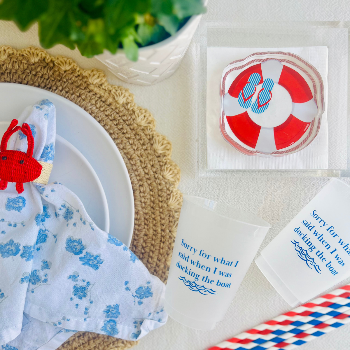 Beach-inspired table setting with a crab napkin ring, frost flex cups that read "Sorry for what I said when I was docking the boat" and  an acrylic napkin weight in the shape of a life buoy is shown from above. 
