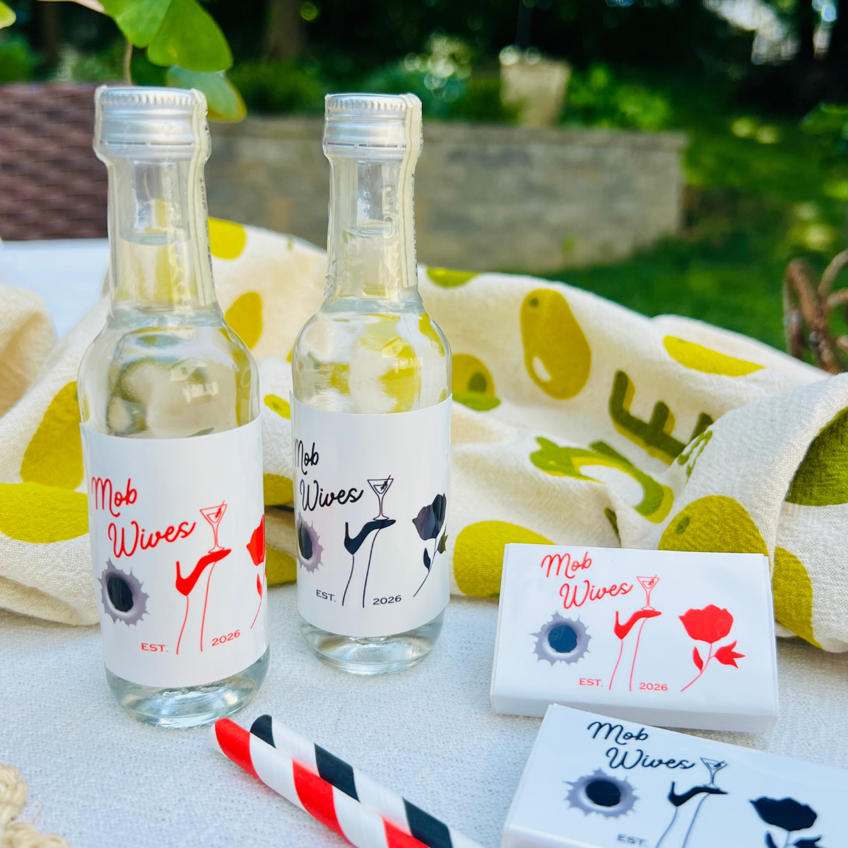 Two mini liquor bottles in a 'Mob Wives' theme show matching, customizable bottle labels and matching matchboxes with the same design are shown on a table in an outdoor setting