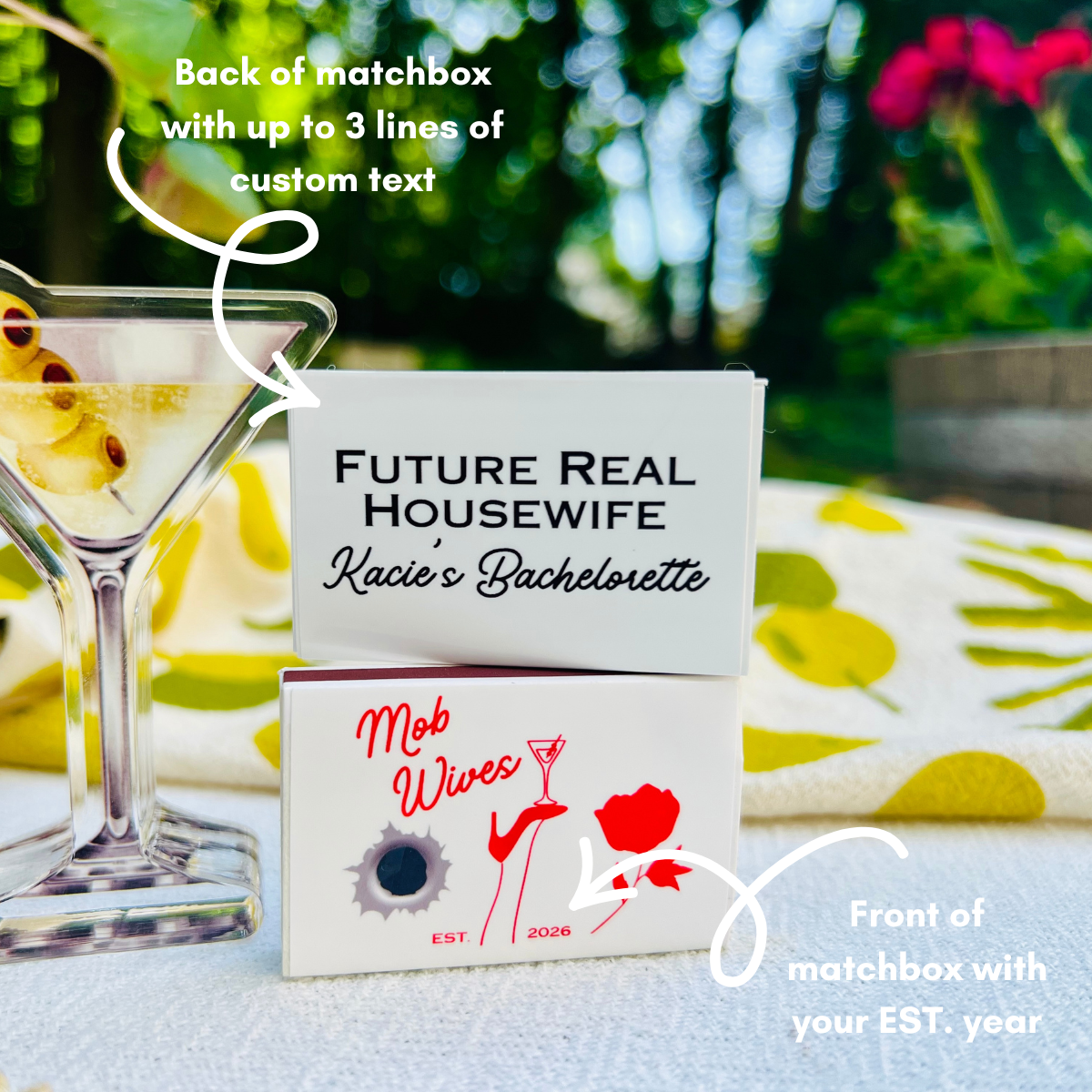 Matchbox with personalized text on a table with a cocktail and garden background. The front and back of the matchboxes are shown, to indicate the different ways these areas can be personalized