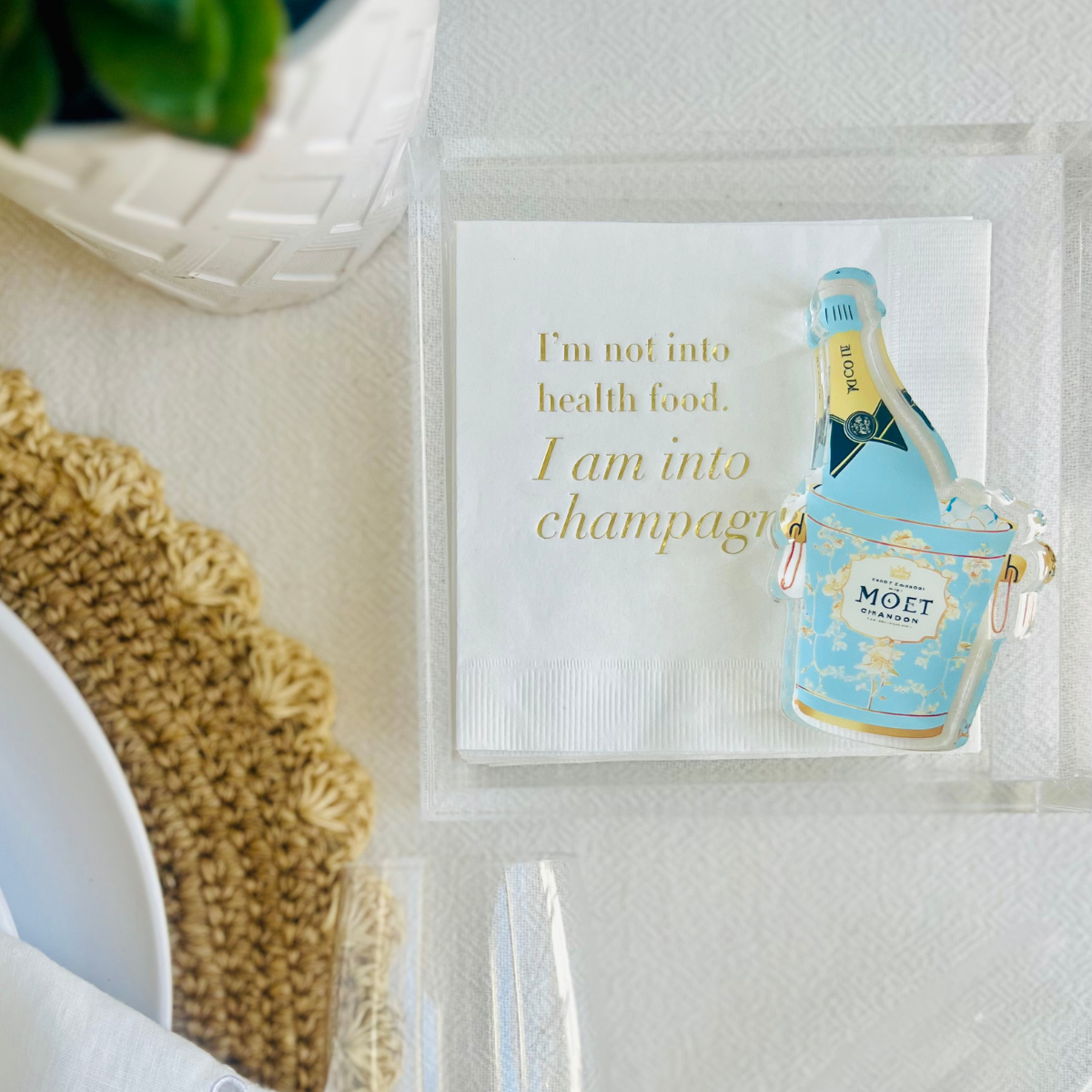 Acrylic napkin weight shaped like a Moet champagne bottle in an ice bucket is shown on top of a stack of white paper napkins that feature funny text that reads "I'm not into health food. I am into champagne."
