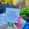 Personalized white cocktail napkin featuring the phrase "New Home, New Baby, New Beginnings" with personalized zip code in elegant matte foil lettering is held up by a hand against a garden background.