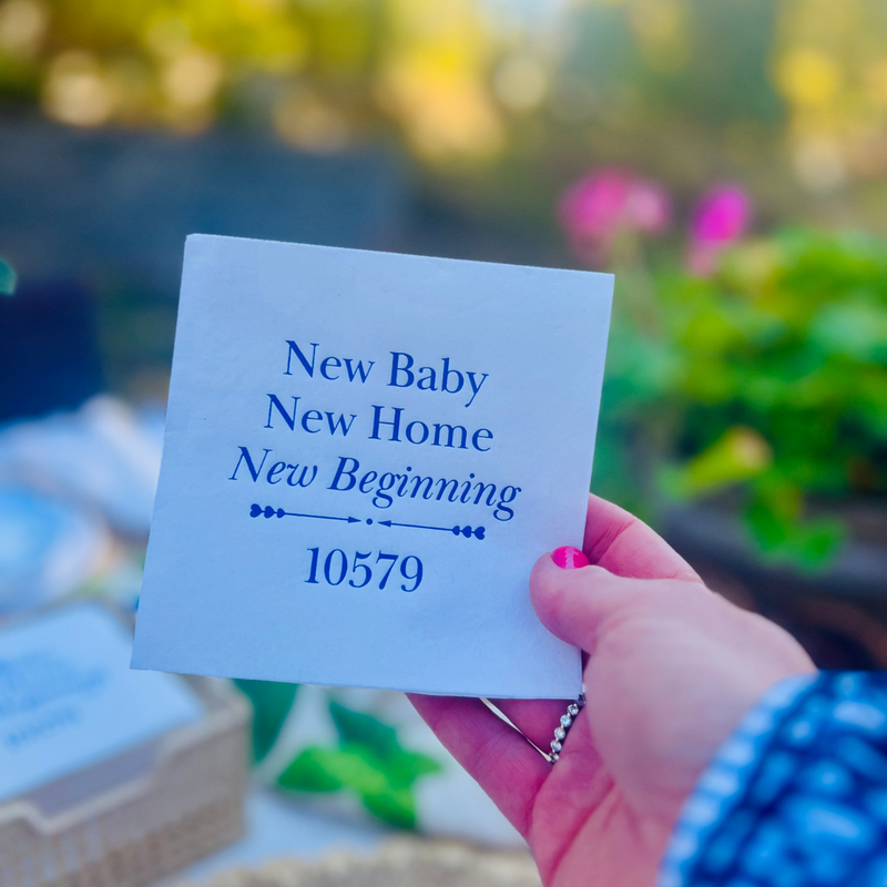 Personalized white cocktail napkin featuring the phrase "New Home, New Baby, New Beginnings" with personalized zip code in elegant matte foil lettering is held up by a hand against a garden background.