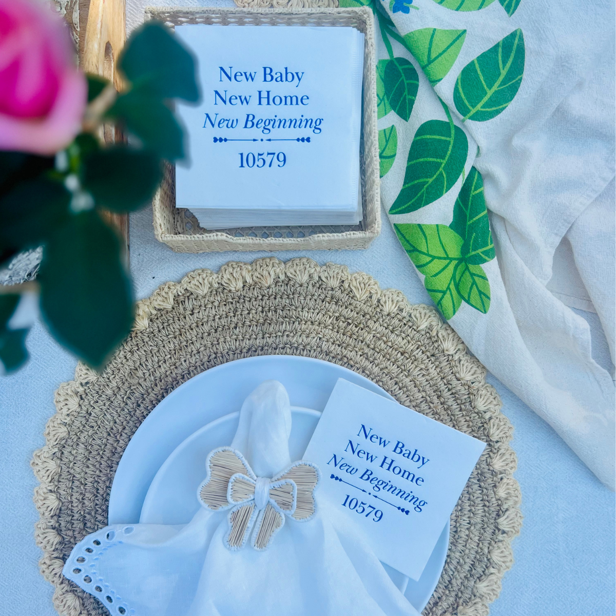 Overhead view of a decorative table setting that shows a linen napkin with a bow napkin ring and a cocktail napkin that reads "New Baby, New Home, New Beginning" tucked behind it. The napkin also features the zip code of the new home - a fun way to celebrate a new chapter. A pink rose and decorative dish towel also appear on the table