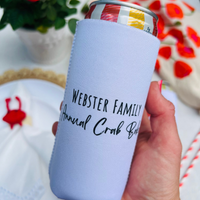White slim can cooler is held above a blurred table. The can cooler shows the back design with text that reads "Webster Family Annual Crab Bake"