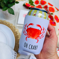 Hand holding a can cooler with a red crab design and 'OH CRAB!' text, blurred background with flowers and plates.