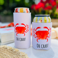 Two can coolers with red crab design and 'OH CRAB!' text on a blurred outdoor background.