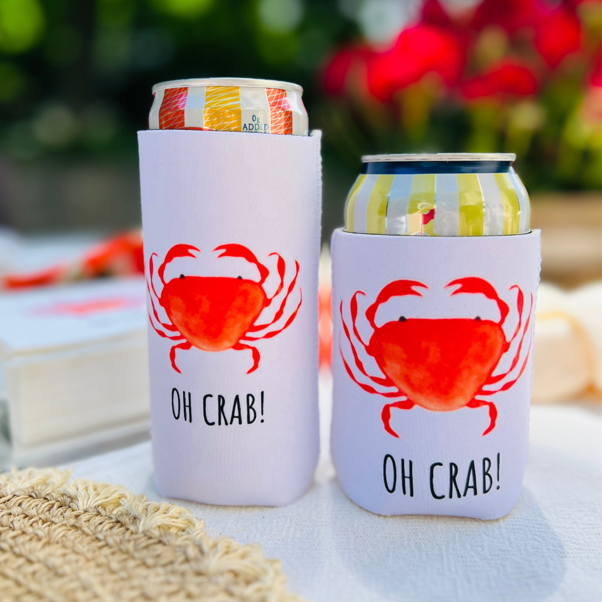 Two can coolers with red crab design and 'OH CRAB!' text on a blurred outdoor background.